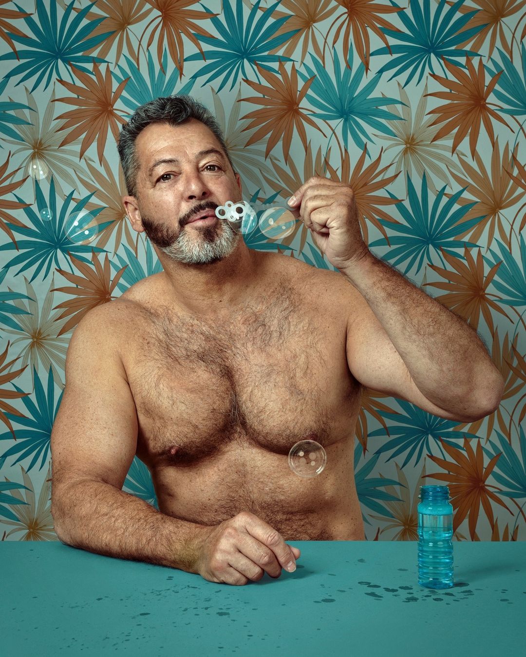 Middle-aged man with a partial beard blows bubbles indoors in front of a teal and brown palm leaf patterned wallpaper, shirtless, eye-level shot.
